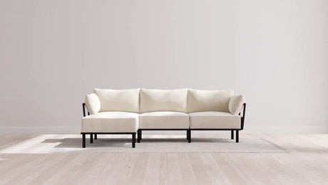A minimalist, cream-colored sectional sofa with black frame on a light-colored rug in a room with pale wooden floors and a plain white wall. Natural light creates soft shadows on the floor.