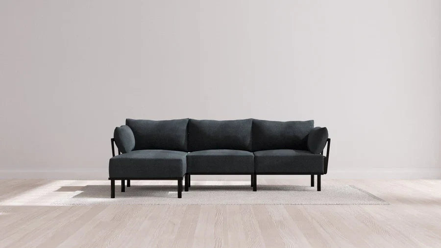 Minimalist gray sectional sofa with black frame positioned on a light-colored area rug. The scene is set against a plain, light gray wall and hardwood floor, creating a simple, modern living room atmosphere.