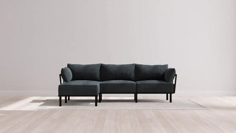 Minimalist gray sectional sofa with black frame positioned on a light-colored area rug. The scene is set against a plain, light gray wall and hardwood floor, creating a simple, modern living room atmosphere.
