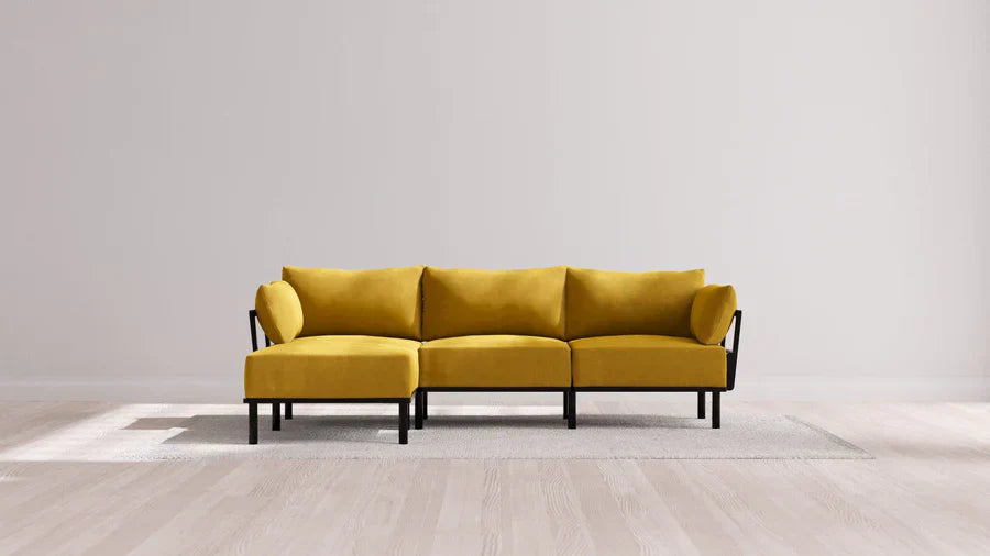 A modern mustard-yellow L-shaped sofa with black metal legs is placed on a light gray rug. The sofa is set against a minimalist white wall on a hardwood floor, creating a clean and contemporary look.