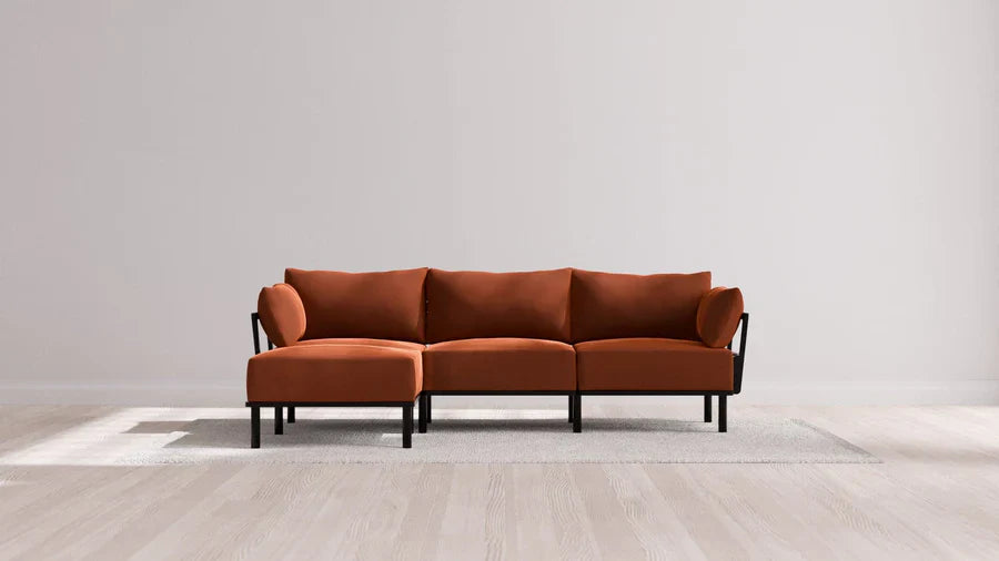 A modern sectional sofa with rust-colored upholstery and black metal frame is centered on a light gray area rug over a wooden floor. The backdrop is a plain, light-colored wall.