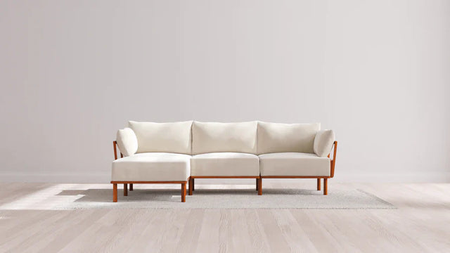 A minimalist living room features a modern white sofa with wooden legs, positioned on a light gray rug. The backdrop is a plain white wall, and the floor is light wood, creating a clean and airy atmosphere.