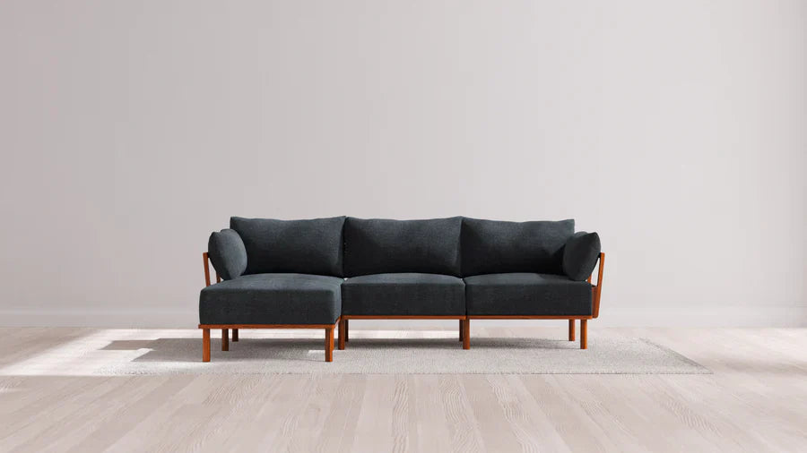 A modern, dark gray sectional sofa with wooden legs is placed on a light-colored rug. The room has a light wood floor and plain white walls, creating a minimalist and contemporary look.