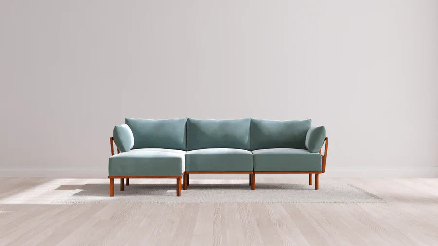 A modern teal sectional sofa with wooden legs is placed on a light-colored rug against a plain white wall in a spacious room with light wood flooring.