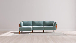 A modern teal sectional sofa with wooden legs is placed on a light-colored rug against a plain white wall in a spacious room with light wood flooring.