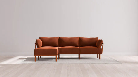 A modern brown sectional sofa with wooden legs is placed on a light gray rug on a wooden floor. The background features a plain white wall, emphasizing the minimalist design of the room.