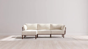 A modern, minimalist white sofa with dark wooden legs is placed on a light-colored rug. The setting is against a plain white wall, and the floor is light wood, creating a clean and spacious look.