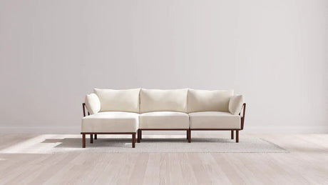 A modern, minimalist white sofa with dark wooden legs is placed on a light-colored rug. The setting is against a plain white wall, and the floor is light wood, creating a clean and spacious look.