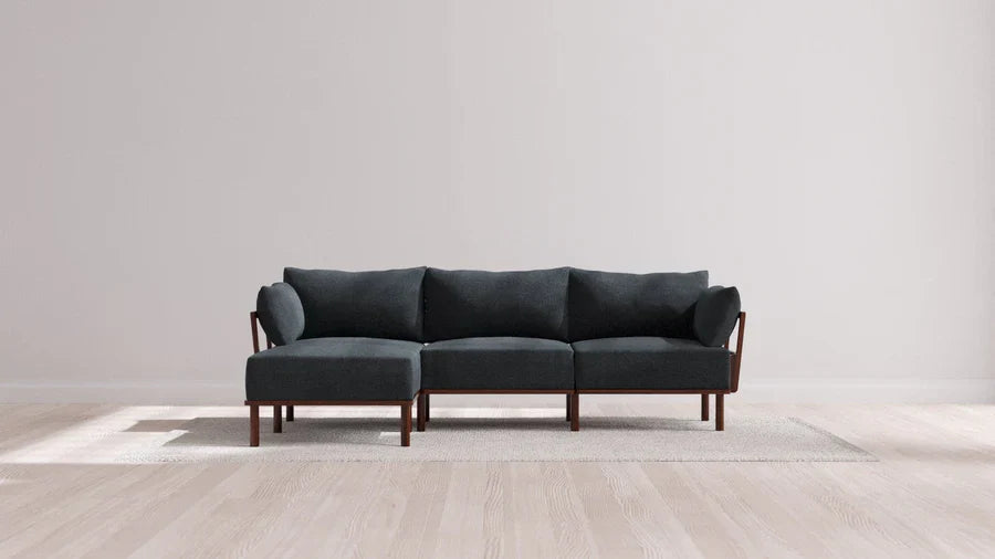 A modern sectional sofa with dark upholstery and wooden legs is placed on a light-colored rug in a minimalistic room with light wood flooring and a plain white wall.