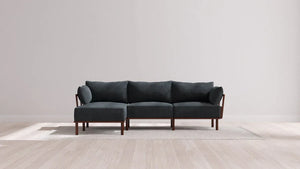 A modern sectional sofa with dark upholstery and wooden legs is placed on a light-colored rug in a minimalistic room with light wood flooring and a plain white wall.