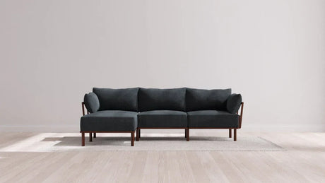 A modern sectional sofa with dark upholstery and wooden legs is placed on a light-colored rug in a minimalistic room with light wood flooring and a plain white wall.