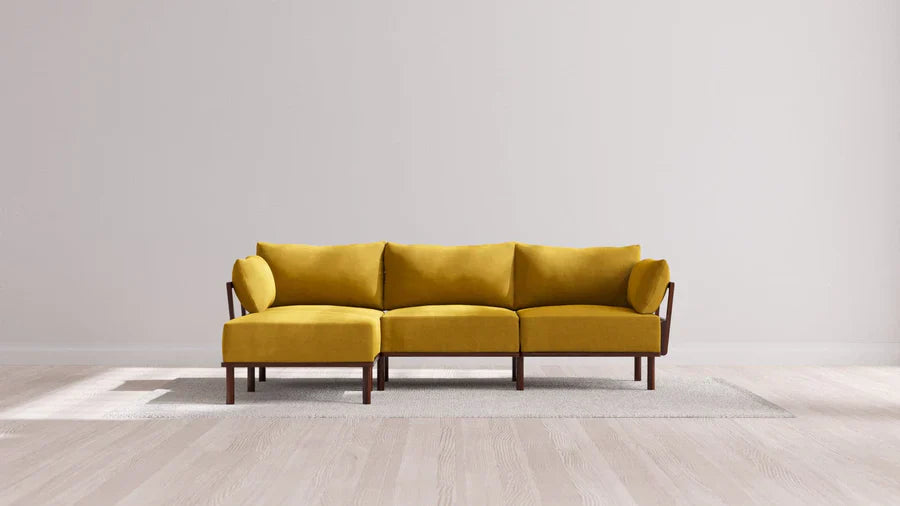 A modern yellow sofa with wooden legs and armrests is positioned against a plain light gray wall. It sits on a light-colored rug on a wooden floor, creating a minimalist and contemporary living space.