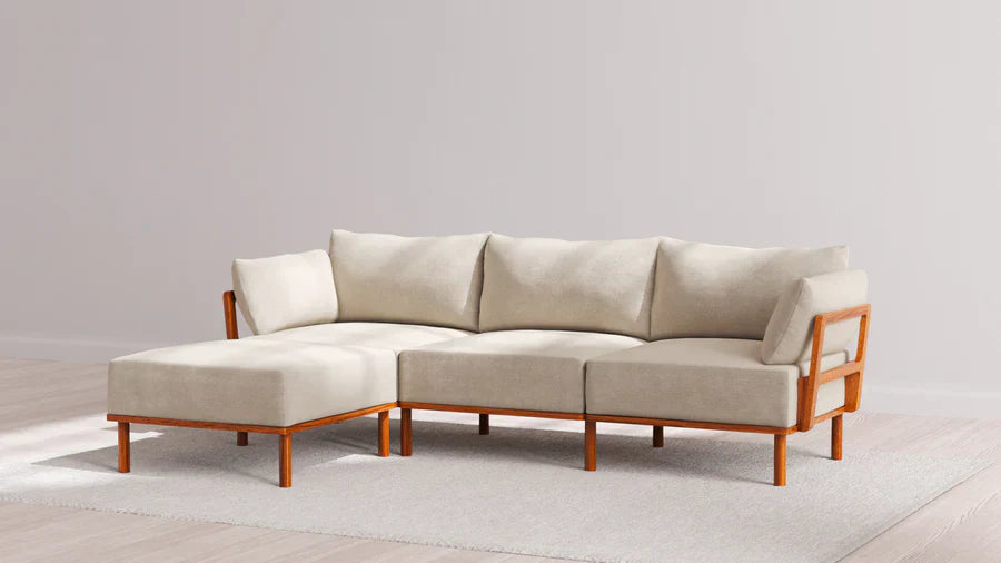 A beige sectional sofa with wooden frame and cushions on a light-colored rug in a minimalist room with wooden flooring and off-white walls.