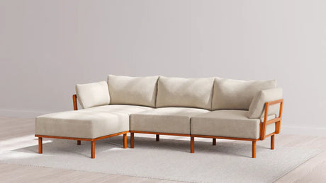 A beige sectional sofa with wooden frame and cushions on a light-colored rug in a minimalist room with wooden flooring and off-white walls.