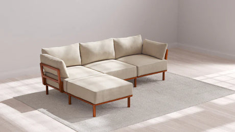A beige sectional sofa with wooden legs is placed on a light gray rug in a bright room with wooden flooring. The sofa has plush cushions and an L-shape design. The setting is minimalistic, with natural light streaming in from the left.