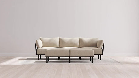 A modern, minimalist living room featuring the Indoor Jack 3-Seater Sofa + Bench Ottoman in Balsa set on a light grey rug. The walls are plain white, and the sunlight casts soft shadows on the wooden floor.