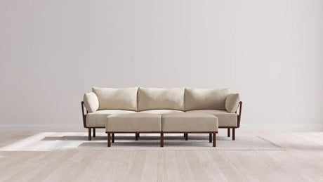 An Indoor Jack 3-Seater Sofa with a Bench Ottoman in Balsa is placed on a light-colored rug in a bright room featuring wooden flooring and plain walls. The sofa's wooden arms and legs enhance the neutral, modern aesthetic.