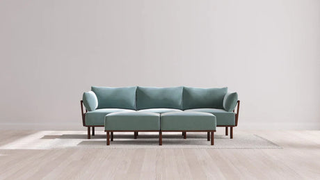 A contemporary living room showcases the Indoor Jack 3-Seater Sofa along with a coordinating Bench Ottoman, both in a soft balsa hue. These pieces feature elegant wooden legs and armrests, resting on a light area rug. The room's decor is minimalist and neutral, boasting a wooden floor and a simple white wall in the background.