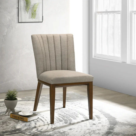 A Stylish Dining Chair from Elmwood, featuring beige upholstery and wooden legs, sits on a patterned rug in a bright room. Nearby on the floor are a small plant and "FAKE BOOKS." A framed fern leaf photo is displayed on the wall near a sunlit window.