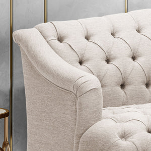 Close-up of the beige, tufted Comfy 3 Seat Sofa with curved armrests, showcasing its fabric texture. A small part of a round wooden side table with a brass rim appears beside the sofa. Minimal background details are shown.