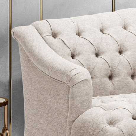 Close-up of the beige, tufted Comfy 3 Seat Sofa with curved armrests, showcasing its fabric texture. A small part of a round wooden side table with a brass rim appears beside the sofa. Minimal background details are shown.