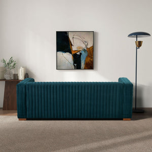 A living room featuring the Elara - Upholstered Sofa in teal, backed by an abstract painting. A modern floor lamp stands on the right, while a wooden table with plants and books sits to the left. The carpeted floor adds warmth to the scene.