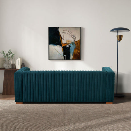 A living room featuring the Elara - Upholstered Sofa in teal, backed by an abstract painting. A modern floor lamp stands on the right, while a wooden table with plants and books sits to the left. The carpeted floor adds warmth to the scene.
