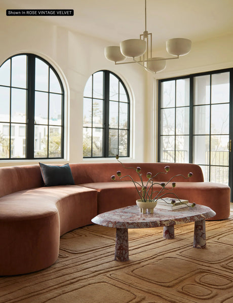 A sunlit living room with large arched windows showcases the Cara Sectional Sofa, a curved piece upholstered in brown velvet. A marble coffee table with a modern vase rests on a textured beige rug, while a stylish white chandelier hangs from the ceiling.