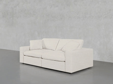 A 2-Seat Modular Loveseat in a modern, light gray finish, featuring two seat cushions and two back cushions, sits against a textured gray wall on a smooth, dark gray floor. The design is minimalist, showcasing a low-profile and rectangular shape.