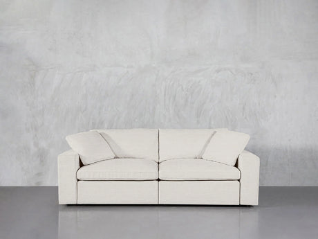 A modern, white 2-Seat Modular Loveseat with seat and back cushions is set against a plain gray, textured wall. The smooth grey floor enhances the minimalist and clean aesthetic.