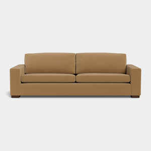 The Barton Sofa, a modern piece with a boxy design, is made from brown leather and showcases two seat cushions and two back cushions made from the same material. It features wide armrests and dark wooden feet, all displayed against a plain white background.