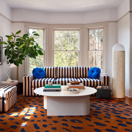 A cozy living room features the Marc Striped Sofa alongside a round table. Blue cushions provide a pop of color, while a tall plant by the bay window brings nature indoors. Books and decor are displayed on the table, and a patterned rug adds warmth to the space.