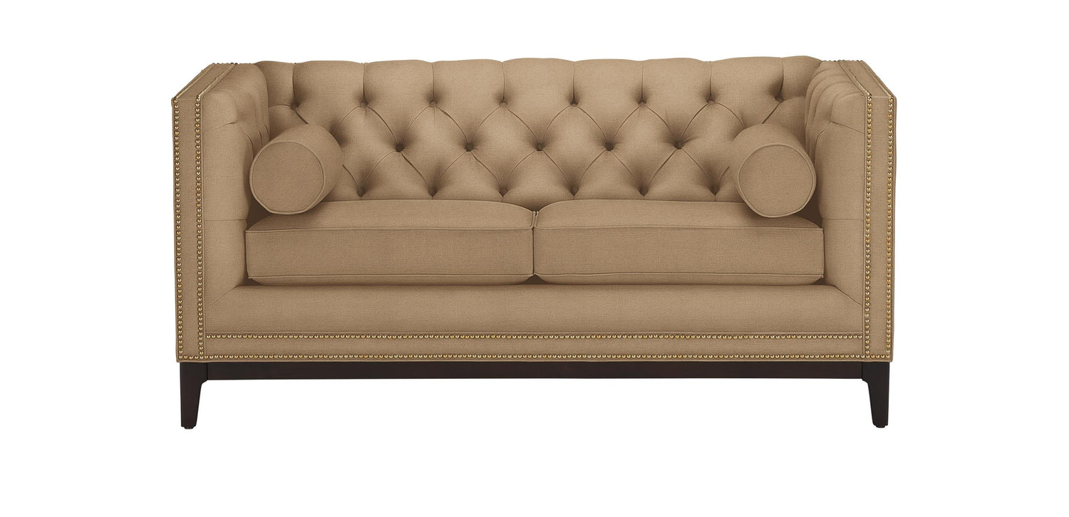 The Anderson Loveseat, in a beige hue with tufted upholstery, is showcased against a white background. It includes two cylindrical pillows and is adorned with nailhead trim along the edges, resting on dark wooden legs.