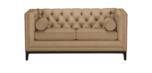 The Anderson Loveseat, in a beige hue with tufted upholstery, is showcased against a white background. It includes two cylindrical pillows and is adorned with nailhead trim along the edges, resting on dark wooden legs.
