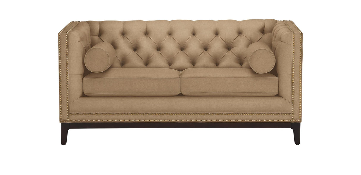 The Anderson Loveseat, in a beige hue with tufted upholstery, is showcased against a white background. It includes two cylindrical pillows and is adorned with nailhead trim along the edges, resting on dark wooden legs.