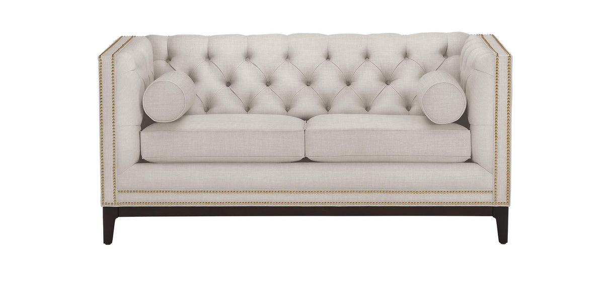 The Anderson Loveseat is a beige tufted seat with a high back and sides, accented by nailhead trim. It features two seat cushions and two cylindrical bolster pillows, supported by dark wooden legs.