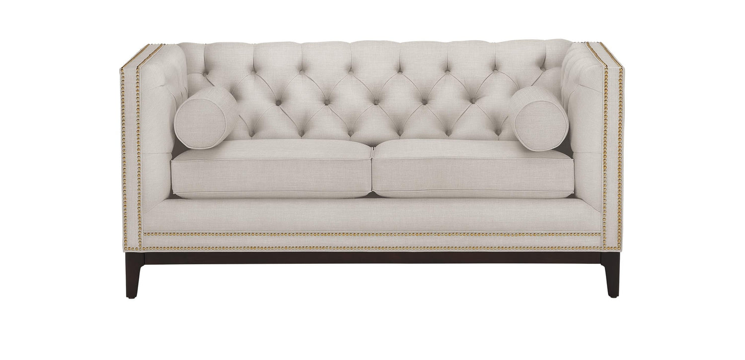 The Anderson Loveseat is a beige tufted seat with a high back and sides, accented by nailhead trim. It features two seat cushions and two cylindrical bolster pillows, supported by dark wooden legs.