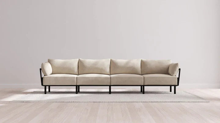 A minimalistic living room showcases the Indoor Jack 4-Seater Sofa in Balsa with black metal legs against a plain wall. The sofa rests on a light-colored rug atop a wooden floor, establishing an elegant and modern atmosphere.