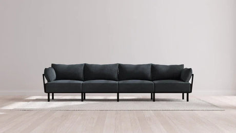 The Indoor Jack 4-Seater Sofa in Balsa, featuring a sleek and modern aesthetic with four cushioned sections and slim armrests, is set on a light gray rug. Its minimalist design is complemented by a plain light-colored wall and a light wood floor.
