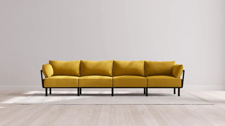 An elongated, minimalist Indoor Jack 4-Seater Sofa in Balsa featuring a yellow hue and four sections rests on a light-colored rug against a neutral wall. The sofa boasts black metal legs and a sleek, modern design, situated on a wooden floor.