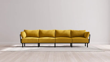 An elongated, minimalist Indoor Jack 4-Seater Sofa in Balsa featuring a yellow hue and four sections rests on a light-colored rug against a neutral wall. The sofa boasts black metal legs and a sleek, modern design, situated on a wooden floor.