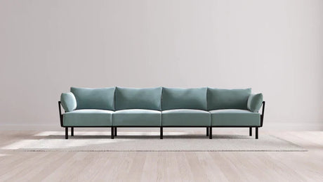 A minimalist living room showcases the Indoor Jack 4-Seater Sofa in Balsa, featuring a long, light teal design with black legs and armrests, placed against a plain white wall. The sofa rests on a light beige rug over a wooden floor.