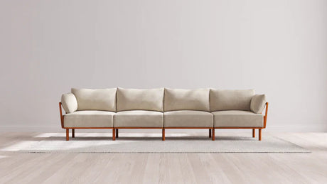 An Indoor Jack 4-Seater Sofa in Balsa, featuring a wooden frame and armrests, sits on a light gray rug in a minimalistic room with light wood flooring and a plain white wall.