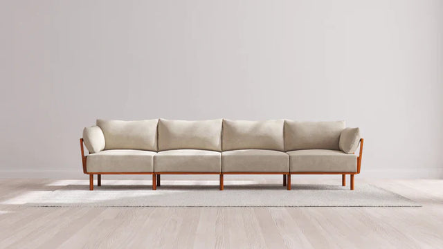 An Indoor Jack 4-Seater Sofa in Balsa, featuring a wooden frame and armrests, sits on a light gray rug in a minimalistic room with light wood flooring and a plain white wall.