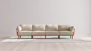 An Indoor Jack 4-Seater Sofa in Balsa, featuring a wooden frame and armrests, sits on a light gray rug in a minimalistic room with light wood flooring and a plain white wall.