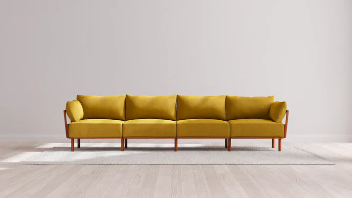 A modern Indoor Jack 4-Seater Sofa in a balsa hue, featuring elegant wooden legs, is centered on a light-colored rug in a minimalist room with a plain white wall and light wood flooring.