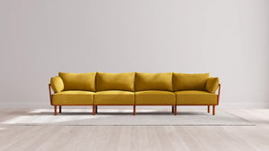A modern Indoor Jack 4-Seater Sofa in a balsa hue, featuring elegant wooden legs, is centered on a light-colored rug in a minimalist room with a plain white wall and light wood flooring.