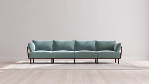 The Indoor Jack 4-Seater Sofa in Balsa, featuring a modern design and light blue upholstery with four sections and wooden legs, is placed on a light gray rug within a minimalist room that boasts a light wood floor and neutral walls.