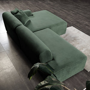 Aerial view of the Mar - High-Quality Sectional Sofa in lush green velvet on a dark tiled floor, with a light gray rug peeking out beneath and large green plant leaves in the corner.