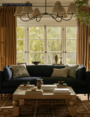 A cozy living room featuring the Fabienne Sofa in dark blue, adorned with patterned cushions. In front of it sits a wooden coffee table holding books and decorative items. Large windows with brown curtains showcase a view of the greenery outside, while a chandelier hangs overhead.